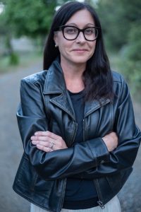 White woman with black hair, black-rimmed glasses, a black leather jacket, and a dark blue/black shirt.